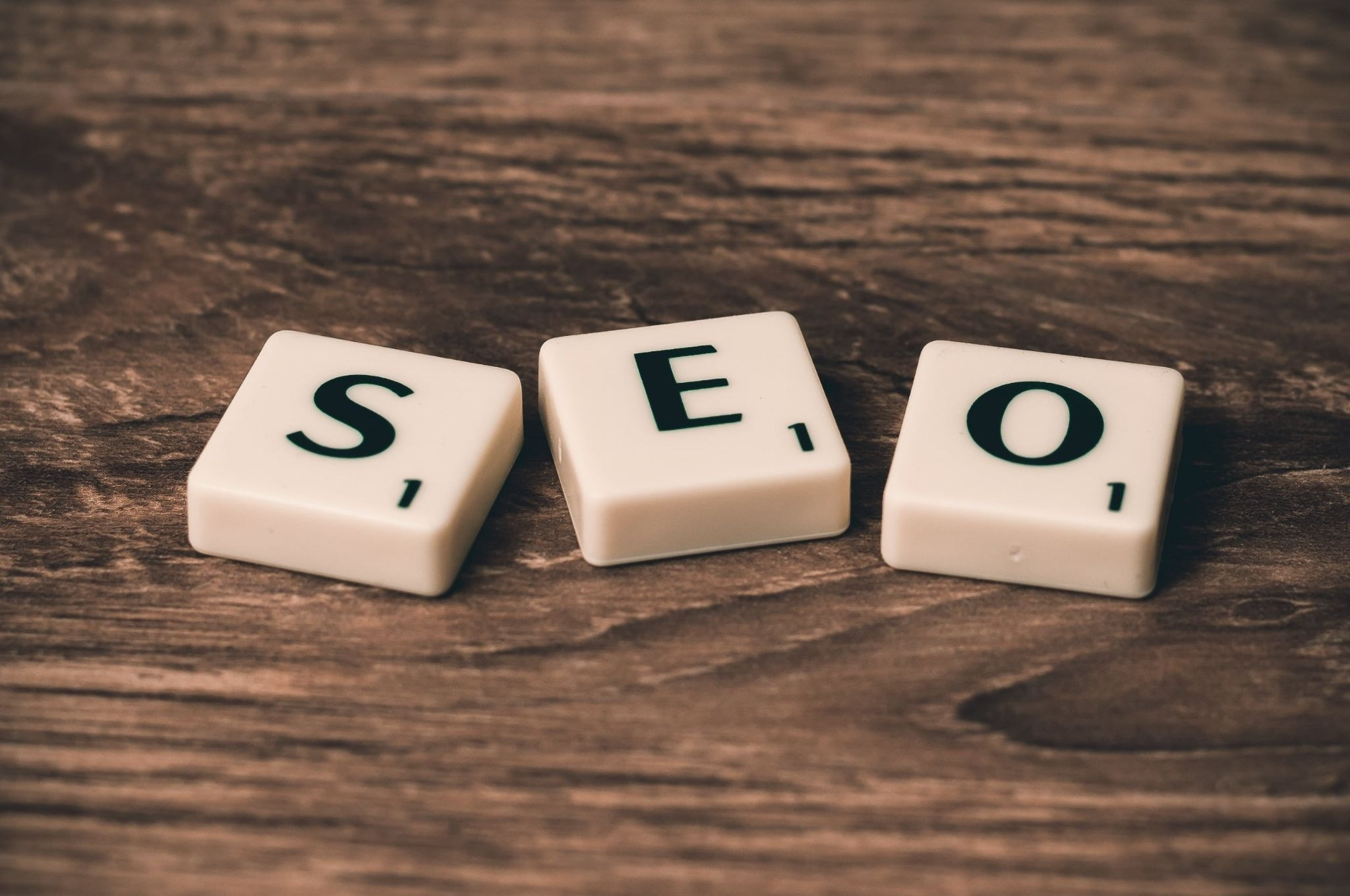 Three white Scrabble tiles spelling "SEO" arranged in a row on a rustic wooden surface, each tile showing one letter — S, E, and O — in bold black font.