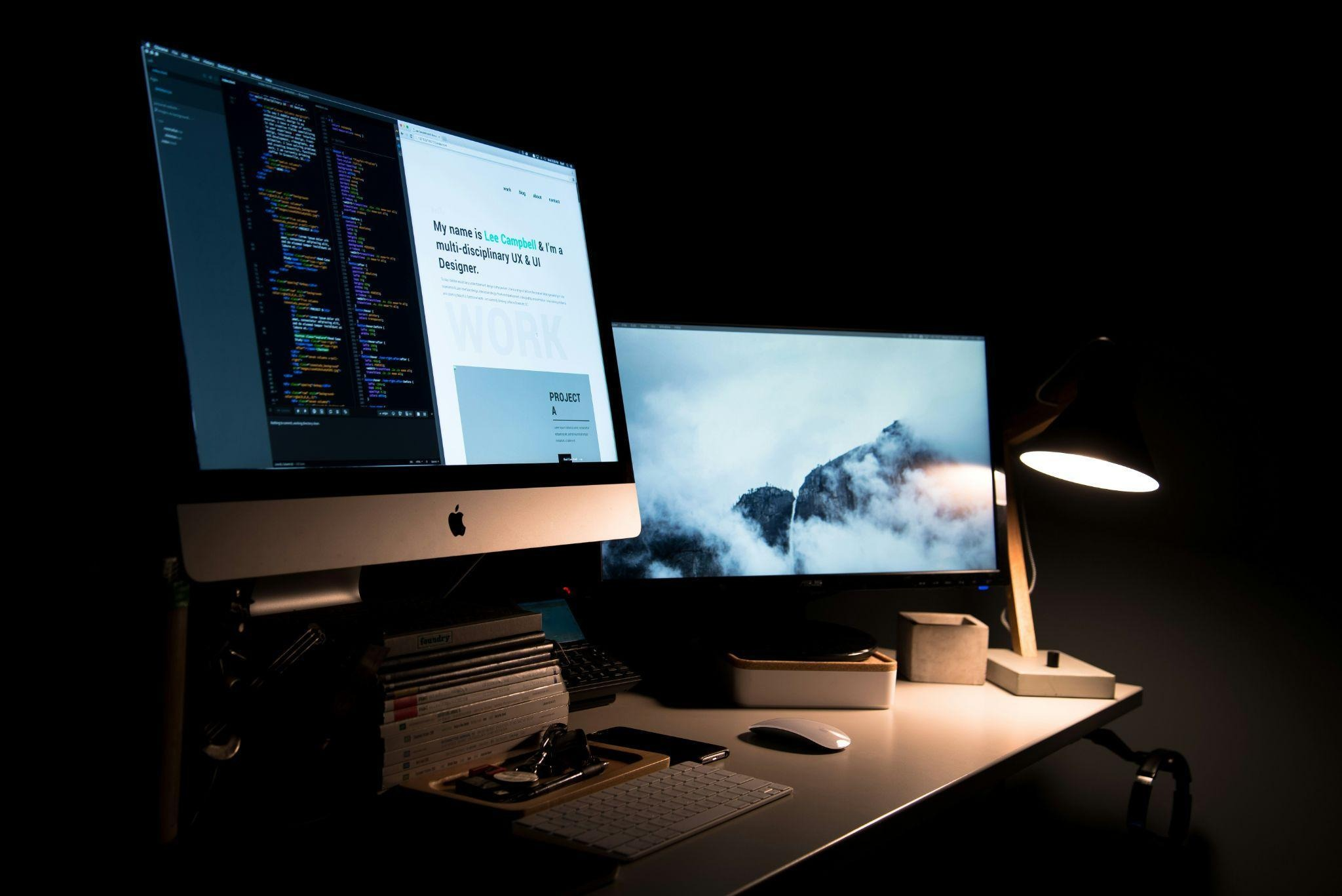 A desk with a dual-monitor setup, a warm desk lamp, a keyboard, a mouse, and a stack of design books. The left monitor shows a code editor next to a portfolio website preview, and the right monitor displays a moody photo of a fog-covered mountain top.