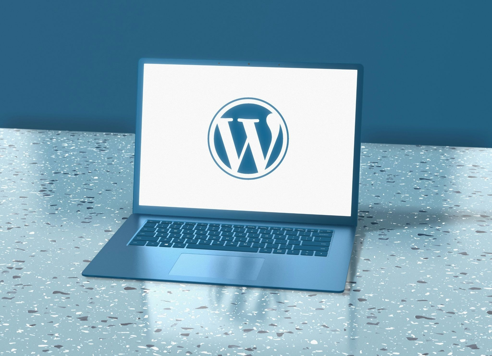 A blue laptop open on a speckled marble surface displaying the WordPress logo on its screen, set against a dark teal blue background.