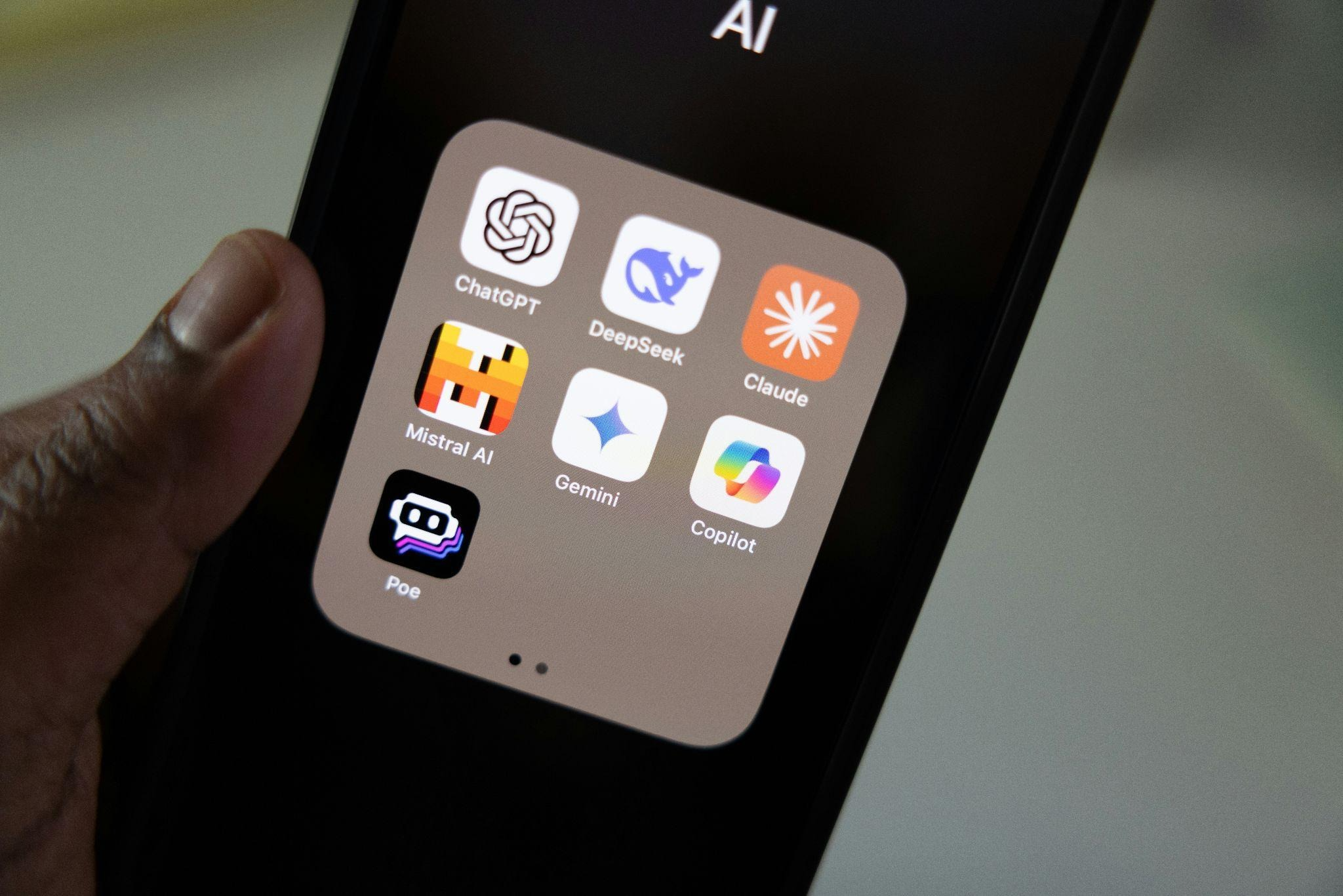 A hand holding a smartphone showing an app folder labeled 'AI' with six AI assistant apps: ChatGPT, DeepSeek, Claude, Mistral AI, Gemini, Copilot, and Poe.