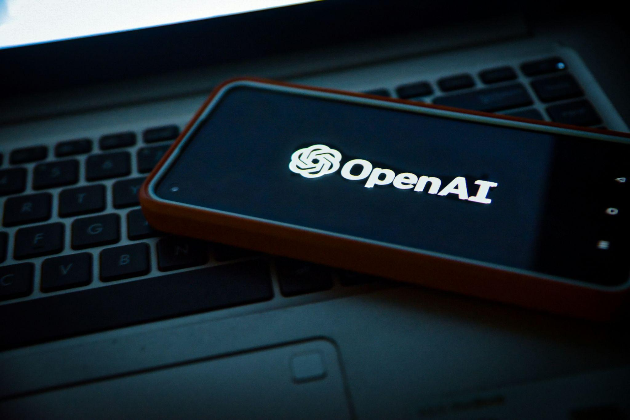 Close-up of a smartphone displaying the OpenAI logo and wordmark on its lock screen, resting on a laptop keyboard in low light.