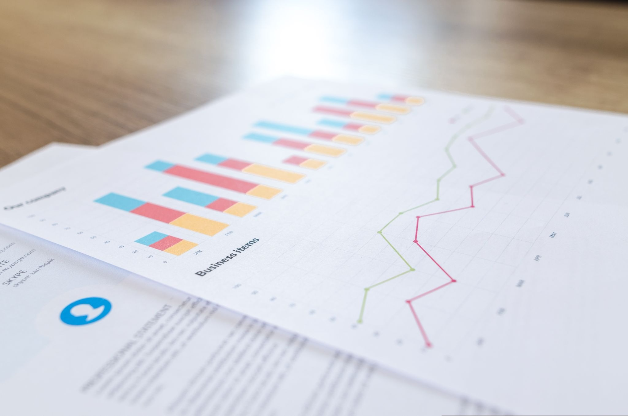Printed business analytics reports laid on a wooden desk, showing colorful grouped bar charts and line graphs tracking performance trends over time, representing website traffic metrics and data visualization.